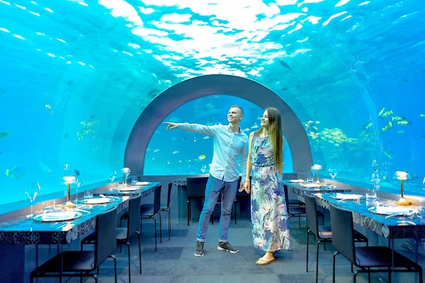 A couple stands inside an underwater restaurant tunnel at You & Me Maldives, set tables and fish visible through the