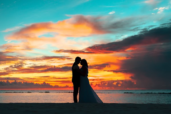 Silhouetted couple embracing on a beach at sunset, bride in a flowing gown, vivid orange and teal sky over the