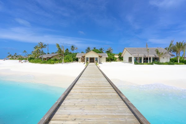 Wooden jetty crossing turquoise water to a white sand beach with thatched-roof buildings and palms at You & Me