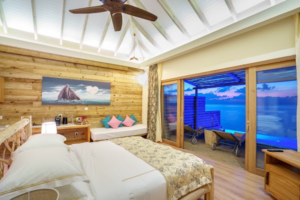 Overwater villa bedroom at You & Me Maldives with wooden interior, white vaulted ceiling and ocean view at dusk through