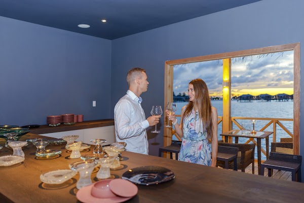 A couple toasting wine glasses at a bar counter with glassware, sunset over the lagoon visible through the open
