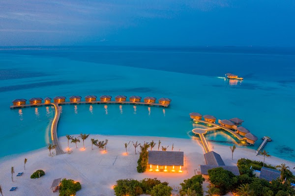Aerial view of overwater villas and lit jetties across a turquoise lagoon at You & Me Maldives during blue