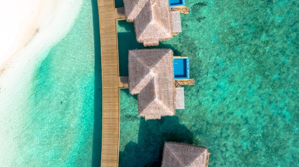 Aerial view of thatched overwater villas beside a wooden jetty in a turquoise lagoon at You & Me Maldives, Raa