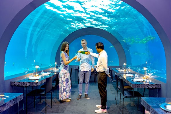 A waiter pours champagne for a couple inside an underwater restaurant with curved glass walls and fish swimming