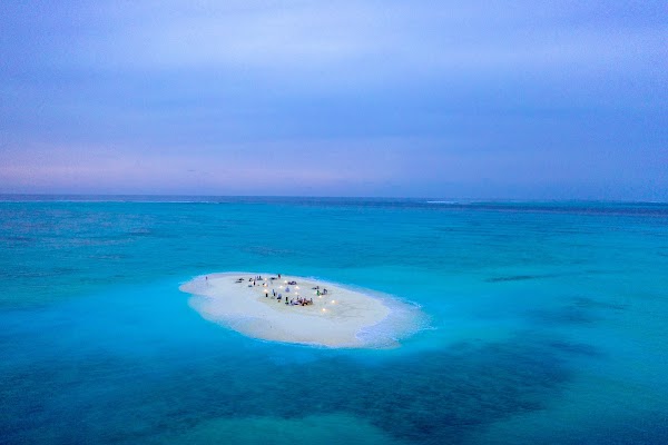 Aerial view of a group gathered around lanterns on a sandbank in a turquoise lagoon at dusk, You & Me
