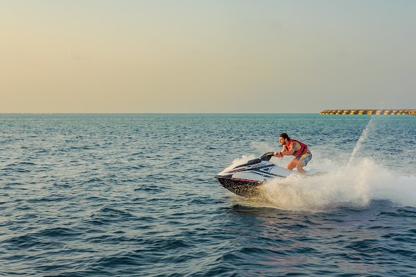 A man in a red life vest rides a jet ski across open sea, spray trailing, overwater villas on the horizon at You & Me