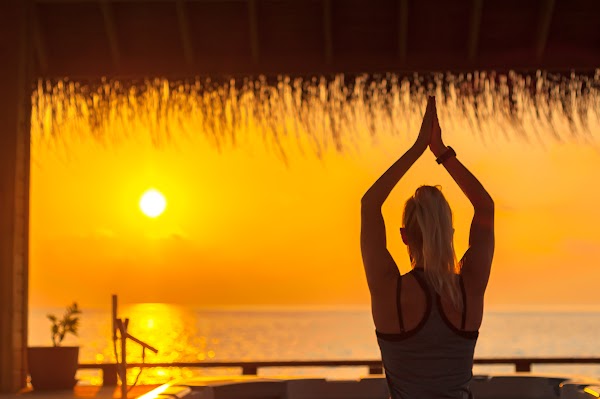 Woman practising yoga with hands raised, silhouetted against a sunset sea at You & Me Maldives, Raa